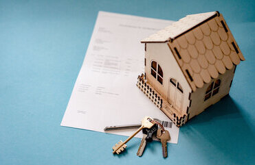 Ein kleines Häusermodell aus Holz ist links im Bild zu sehen. darunter liegt ein Dokument und darauf ein Schlüsselbund.