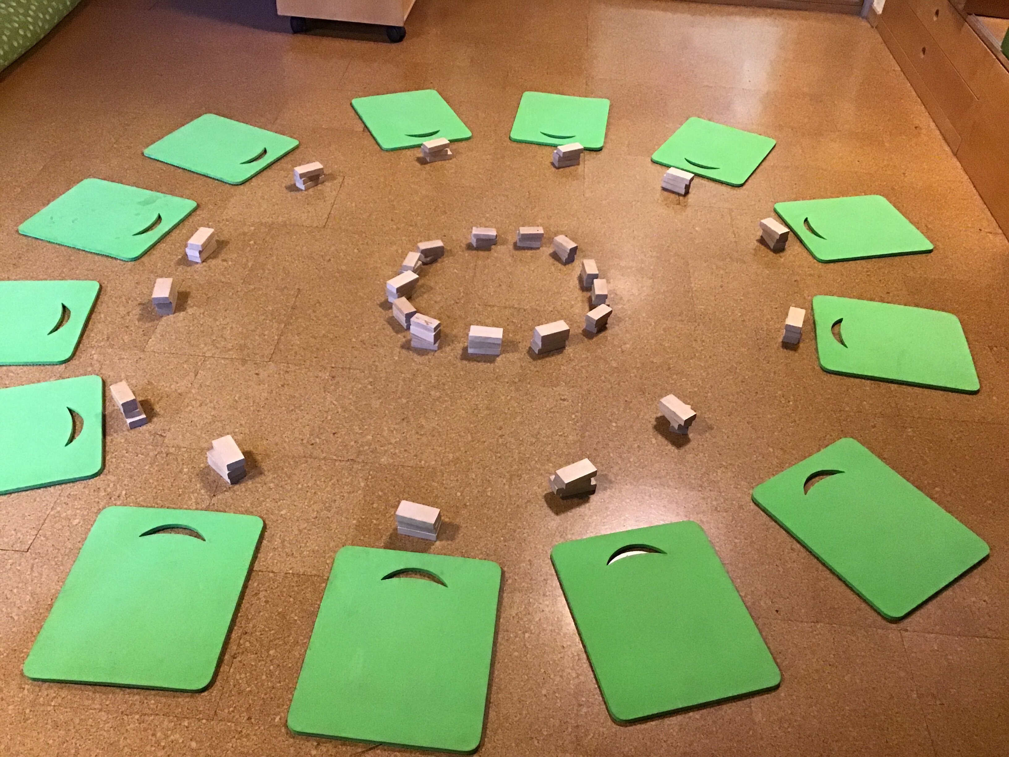 In einem Kreis liegen auf dem Boden dreizehn grüne viereckige Sitzkissen. Davor befindet sich jeweils ein Bauklotz. In der Mitte ist ein kleiner Kreis aus Bauklötzern aufgestellt.