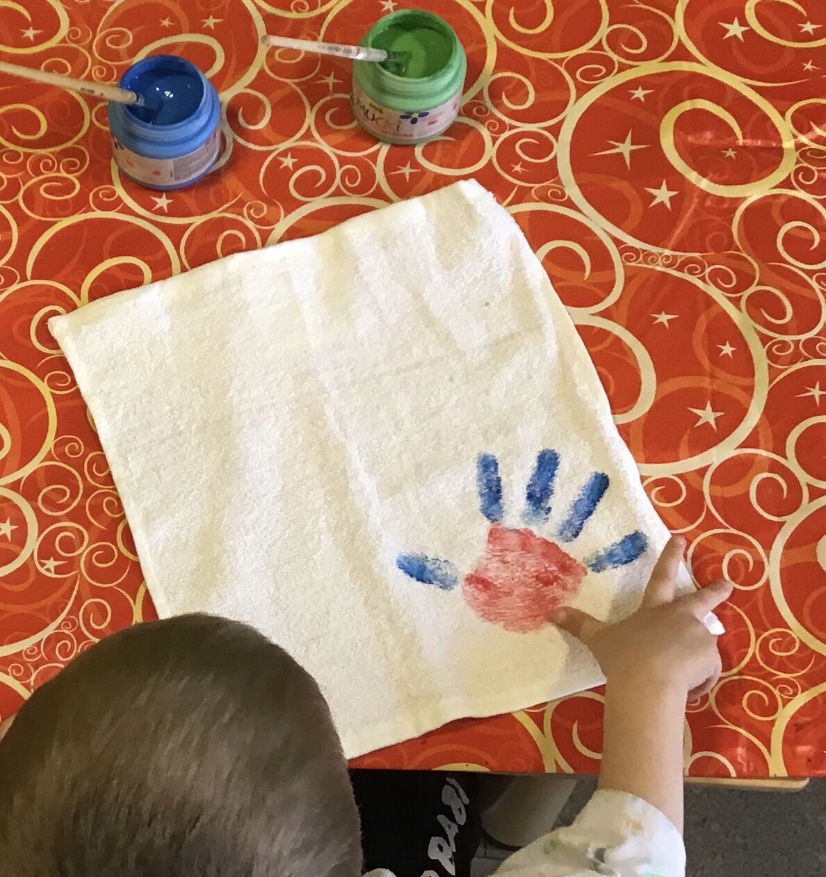 Von oben sieht man den Hinterkopf eines Kindes im linken Bildrand. Das Kind hat gerade einen blau - roten Handabdruck auf ein Stück weissen Stoff vor ihm aufgebracht.